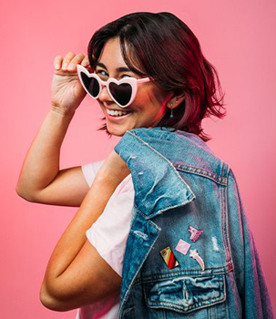 woman wearing pink heart-shaped sunglasses and carrying a jean jacket with enamel pins